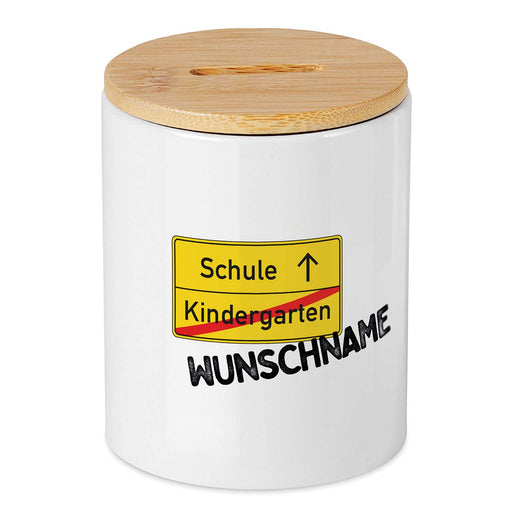 Keramikspardose mit Bambusdeckel zum Öffnen in weiß mit Ortsschild Kindergarten durchgestrichen und Schule mit Pfeil nach oben und Wunschname